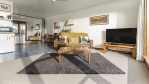 Una sala de estar moderna con un sofá, una mesa de centro y un televisor. Se observa un área de cocina al fondo.
