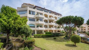 Un hotel de varios pisos con balcones, un pequeño jardín con un estanque y vegetación circundante.