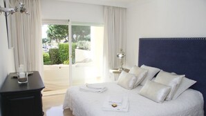 Una cama bien hecha con almohadas y una toalla doblada, una lámpara de noche y vistas a un espacio exterior verde a través de una puerta corredera.