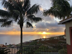 A balcony with a view of a sunset over the sea, palm trees, and houses on a hillside. - Traditional house. Fantastic sea view. Large outdoor spaces with barbecue. (Calheta)