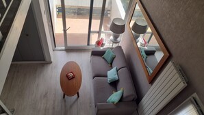 A modern living room with a grey sofa, a wooden coffee table, and a large window offering a view of a balcony. - Spacious T2 duplex in a wooded residence 5 minutes from the beach and the center (Andernos-les-Bains)