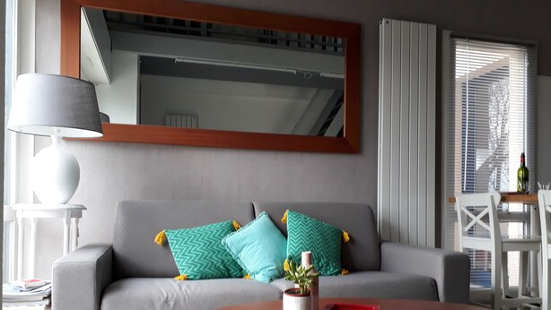A modern living room with a grey sofa, two patterned cushions, a round wooden coffee table, a white side table with a lamp, and a dining area with a table and chairs. - Spacious T2 duplex in a wooded residence 5 minutes from the beach and the center (Andernos-les-Bains)
