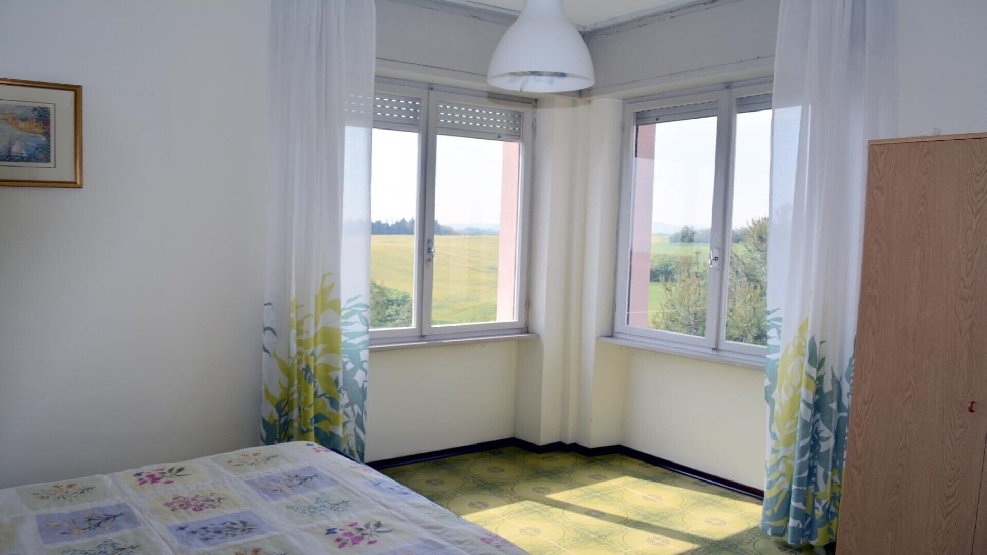 A bedroom with two windows, a bed with floral bedding, and a view of a green landscape.