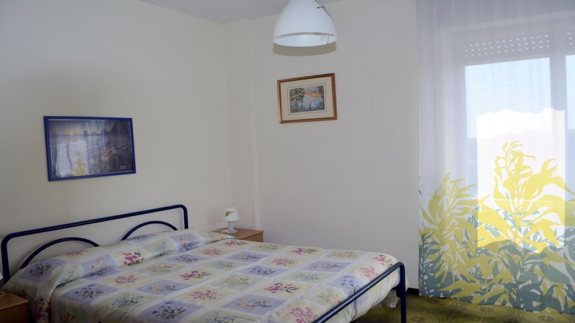 Ein Schlafzimmer mit einem Bett, einem Nachttisch, einer Lampe, einem gerahmten Bild und einem Fenster mit Blick.
