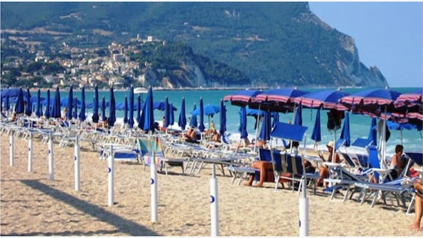 Una spiaggia con file di lettini e ombrelloni, vista libera sul mare e montagne sullo sfondo.