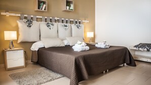 Une chambre d’hôtel bien aménagée, avec un grand lit, deux tables de chevet, une petite table d’appoint, une petite étagère avec des livres et une plante, et une lampe de chevet.