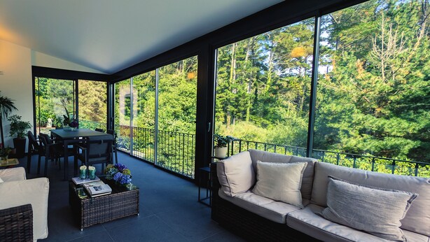 Ein modernes Interieur mit einem großen Fenster mit Blick auf einen Wald. Es gibt eine Essecke mit Tisch und Stühlen, ein Sofa und einen kleinen Tisch mit Getränken und Blumen.