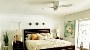Quarto com cama, mesas de cabeceira, ventilador de teto e uma janela grande.