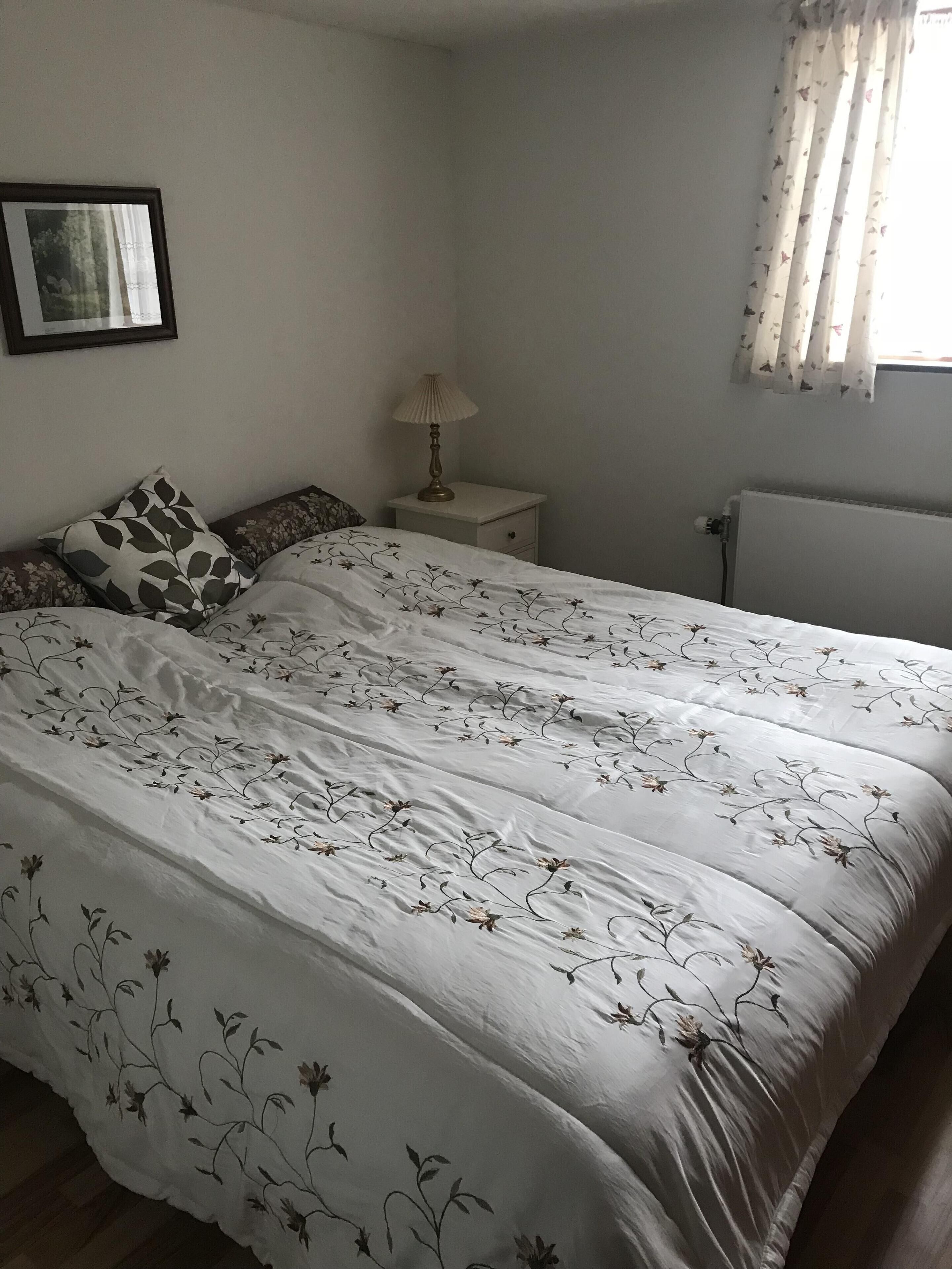 Un dormitorio con una cama, una mesita de noche, una lámpara, un cuadro enmarcado en la pared y una ventana con cortinas.