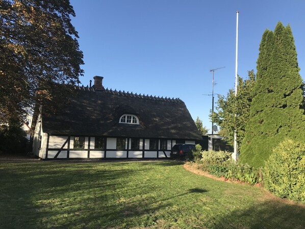 Una casa tradicional con techo de paja, una ventana pequeña y una chimenea, rodeada por un césped cuidado y árboles altos.