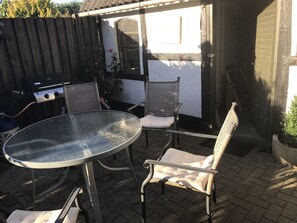 Un patio con una mesa redonda de vidrio y dos sillas de mimbre con cojines, una parrilla y un edificio blanco al fondo.