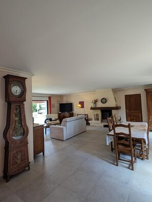 Un salon spacieux avec une cheminée, une grande horloge, un téléviseur et un coin repas avec des chaises en bois.