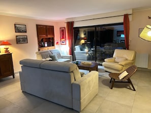 Un salon comprenant un canapé, des fauteuils, une table basse et un coin repas visible à travers une porte vitrée.