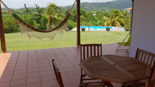 Un patio con una mesa y sillas de madera, una hamaca y vistas a una piscina y abundante vegetación.