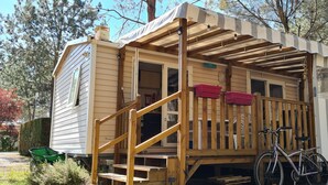 A mobile home with a wooden porch, a bicycle, and a small outdoor seating area. - Mobile home in the Landes, view of the forest (Saint-Julien-en-Born)