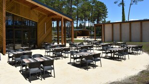 An outdoor patio with multiple tables and chairs, surrounded by a wooden building with large windows. - Mobile home in the Landes, view of the forest (Saint-Julien-en-Born)