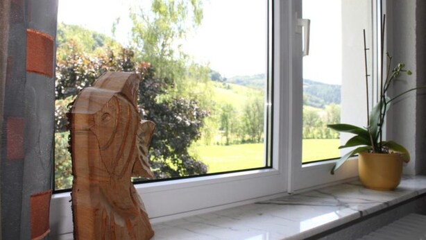 Une sculpture en bois, posée sur un rebord de fenêtre, d’où l’on peut admirer un paysage verdoyant.