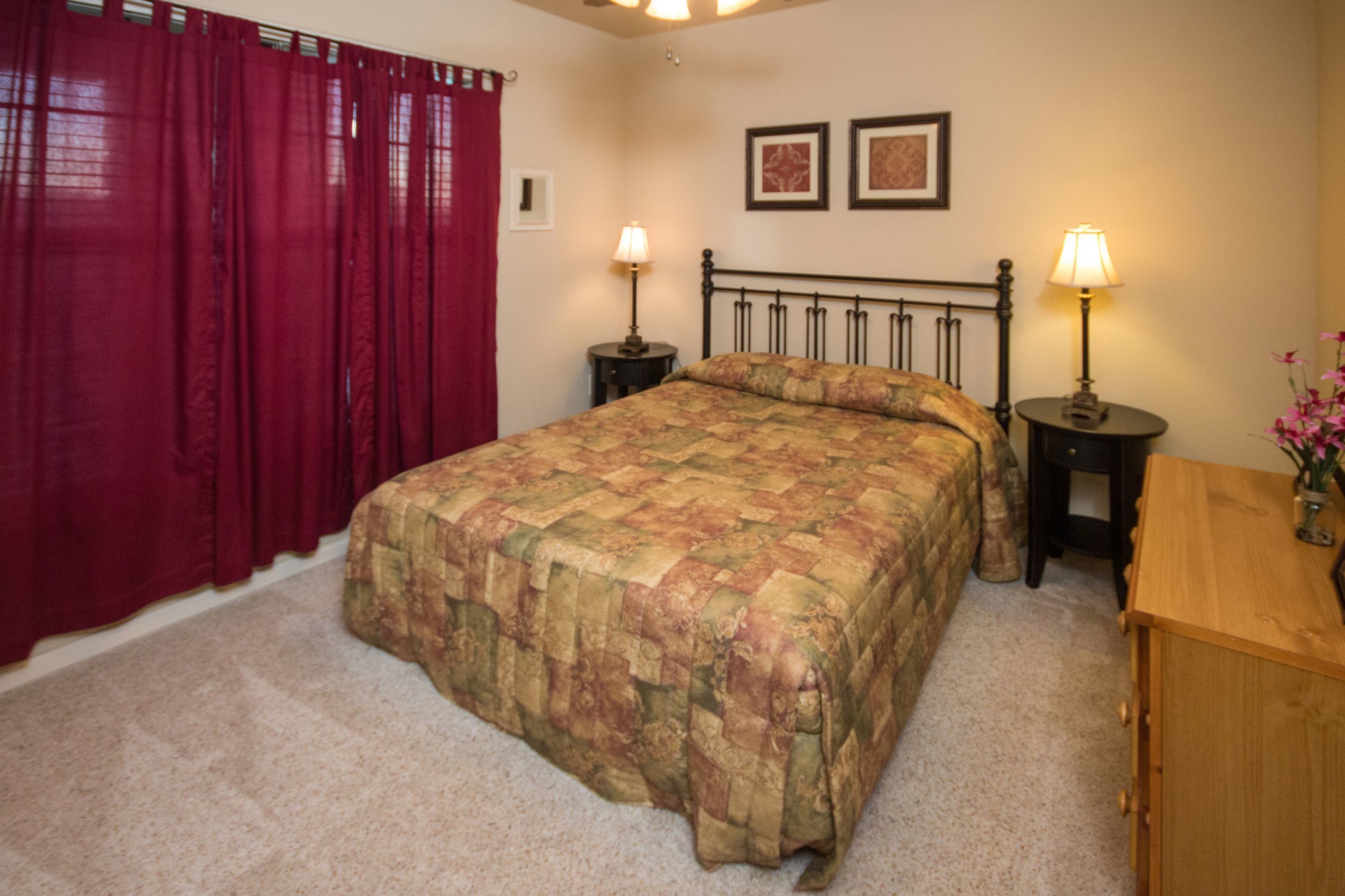 A bedroom with a bed, two bedside tables with lamps, a wooden nightstand, a red curtain, and two framed pictures on the wall.