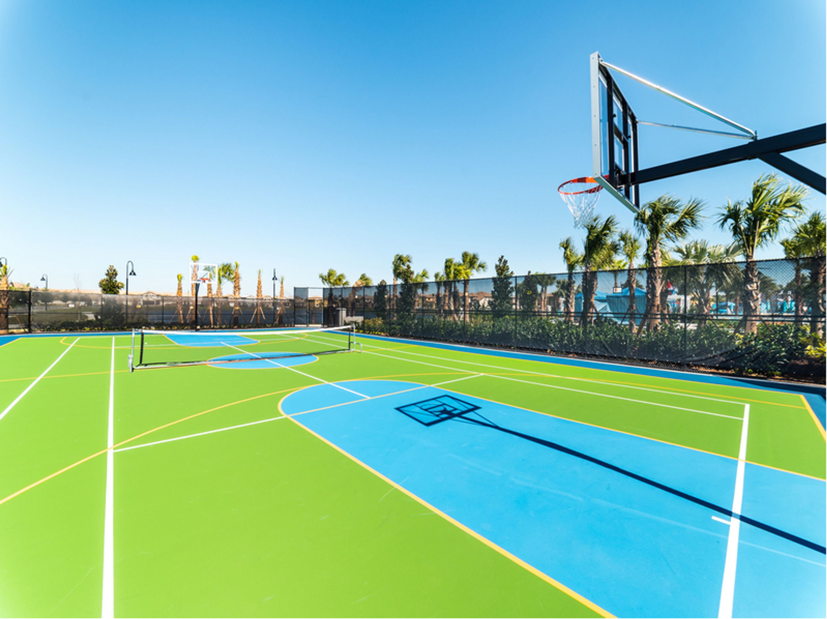 An outdoor basketball court with a clear sky, palm trees, and a fence.