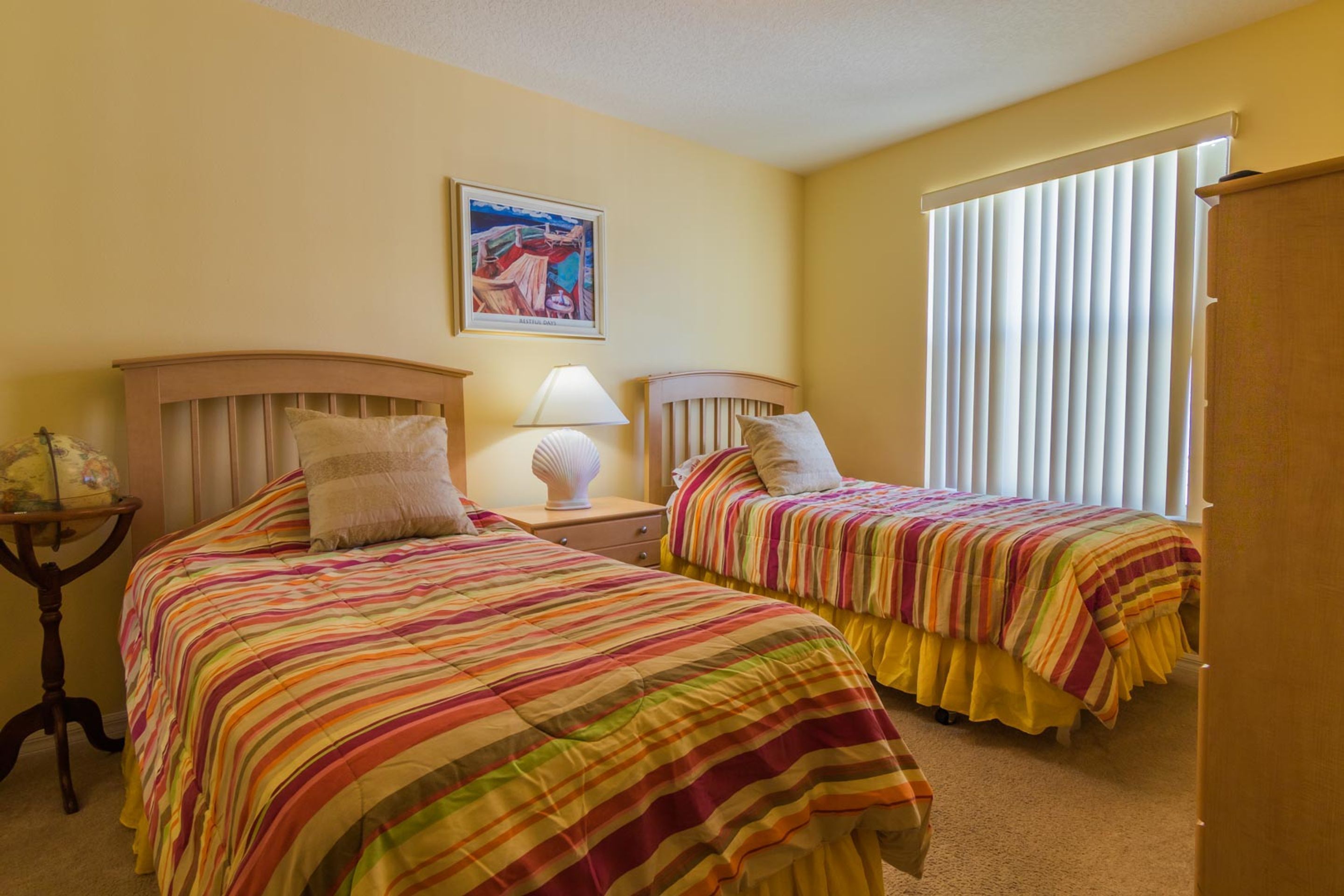 A room with two beds, each with a striped bedspread, a bedside table with a lamp, a framed picture on the wall, and a window with blinds.