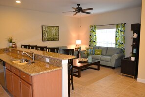 A modern kitchen with granite countertops, a dining area with a gray sofa, and a ceiling fan.
