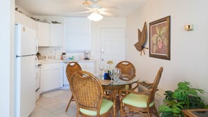 A compact kitchen with white cabinets, a dining table with wicker chairs, a ceiling fan, and a door with a framed picture. - Bonita Beach 5500 - 1906 (Bonita Springs)