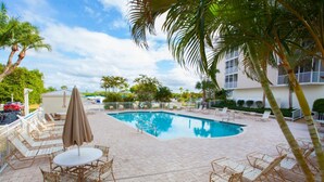 A pool area with lounge chairs and umbrellas, surrounded by palm trees and a building in the background. - Bonita Beach 5500 - 1906 (Bonita Springs)