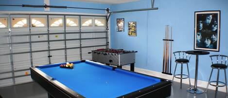 A pool table with a blue felt surface, a foosball table, and two bar stools in a room with blue walls and a garage door.
