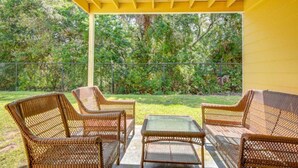 A covered patio area with wicker furniture and a glass table, surrounded by a yellow wall and a view of a lush garden.