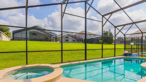 Una piscina amb un banyera d'hidromassatge, envoltada per una tanca metàl·lica i una zona verda amb cases al fons.