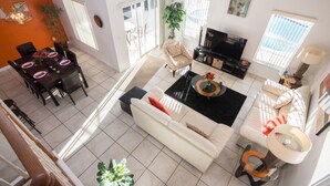 Un salón espacioso con un sofá blanco, una mesa de centro negra y un televisor. Hay una zona de comedor con una mesa y sillas, y un balcón con una silla.