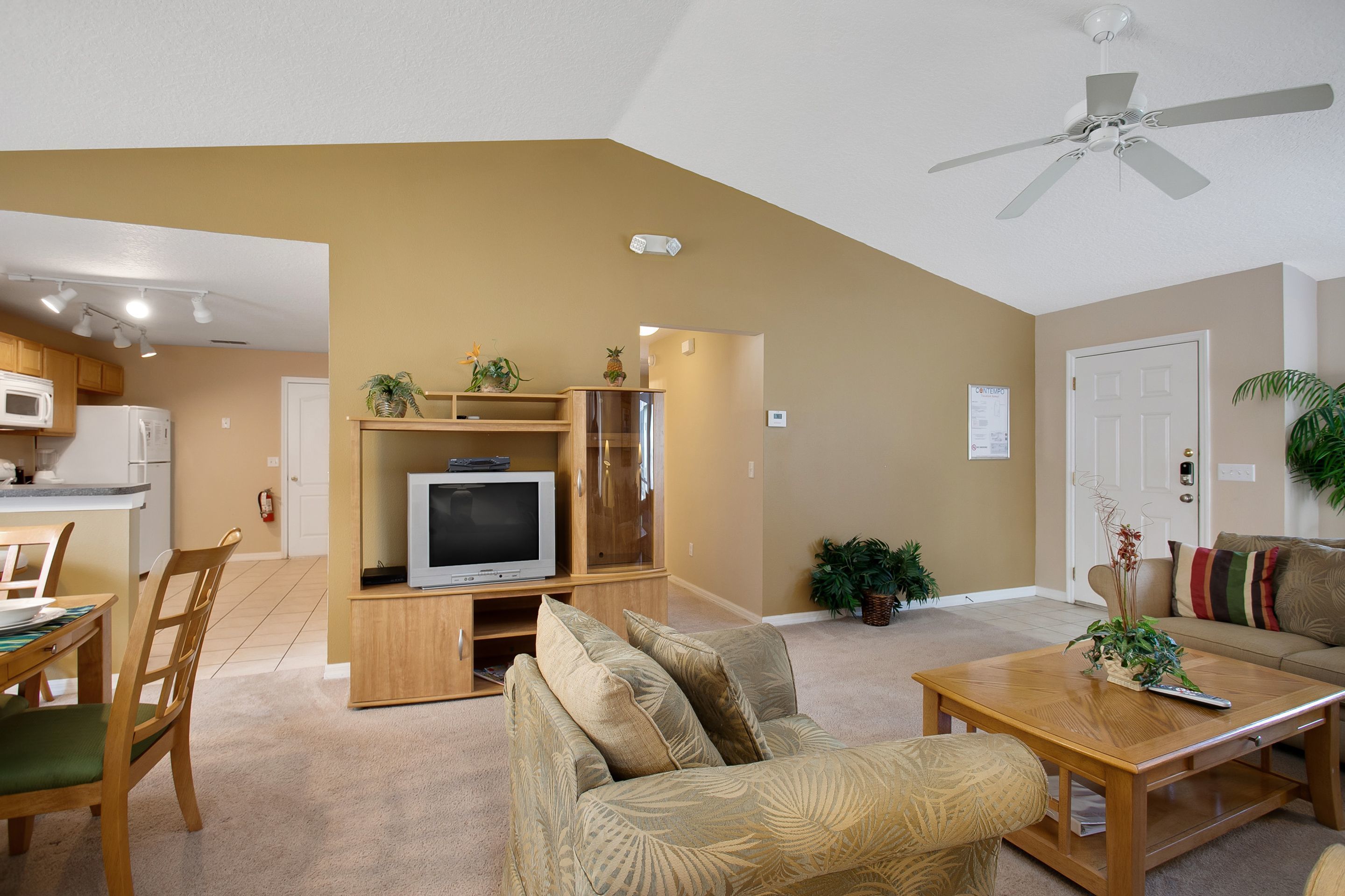 A living room with a sofa, coffee table, and TV stand. There is a kitchen area with a microwave and cabinets. The room has a ceiling fan and a door.