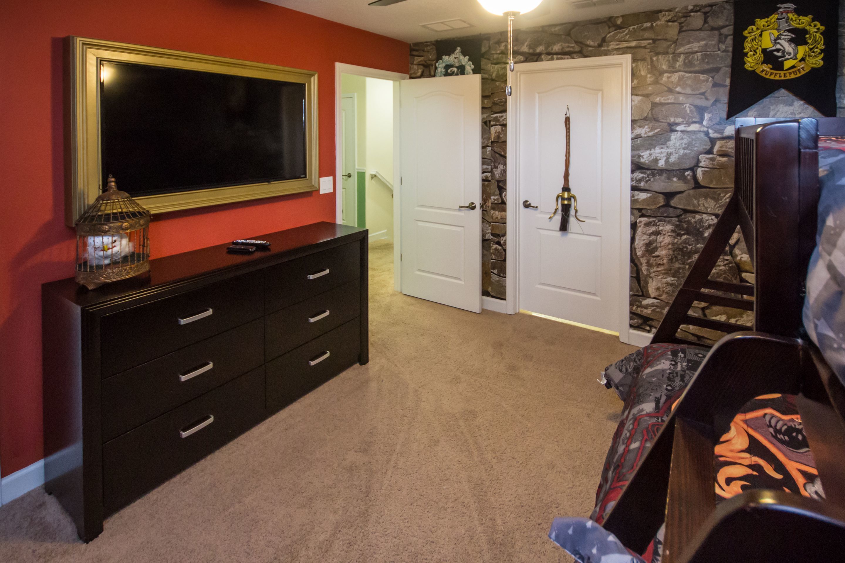 A room with a large flat-screen TV mounted on a wall, a dark wooden dresser with drawers, a stone accent wall, and a door with a brass handle.