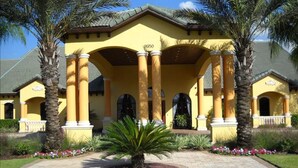 A yellow building with columns and arched doorways, surrounded by palm trees and a paved walkway. - Paradise Palms 8959 (Kissimmee)