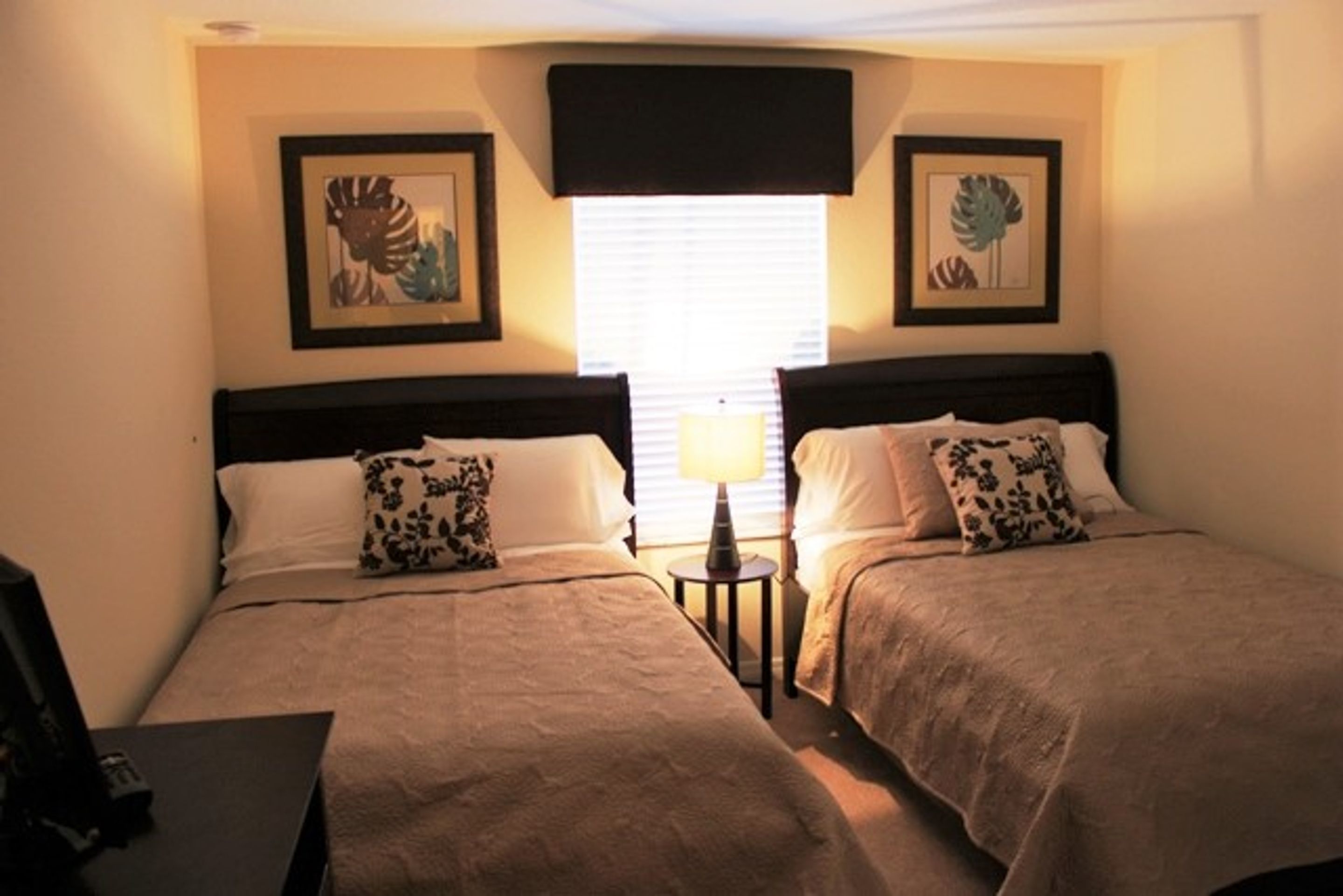 A hotel room with two beds, each with a pillow and a bedside table with a lamp. There are framed pictures on the wall above the beds.