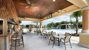 A covered outdoor area with tables and chairs, a pool, and a TV screen.