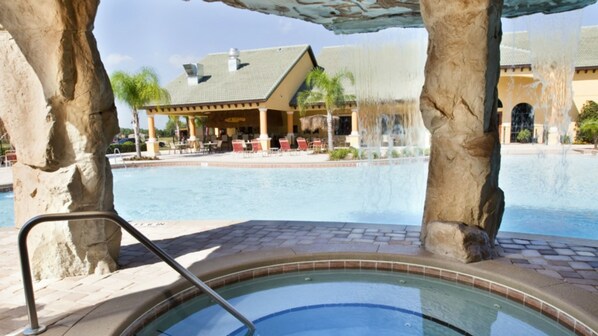 A pool area with a hot tub, surrounded by stone pillars, a covered seating area, and a building with a tiled roof in the background.