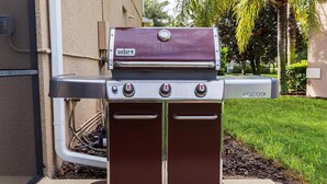 A Weber Genesis portable grill with three burners, a digital display, and a stainless steel finish, positioned on a concrete pad in a residential setting.