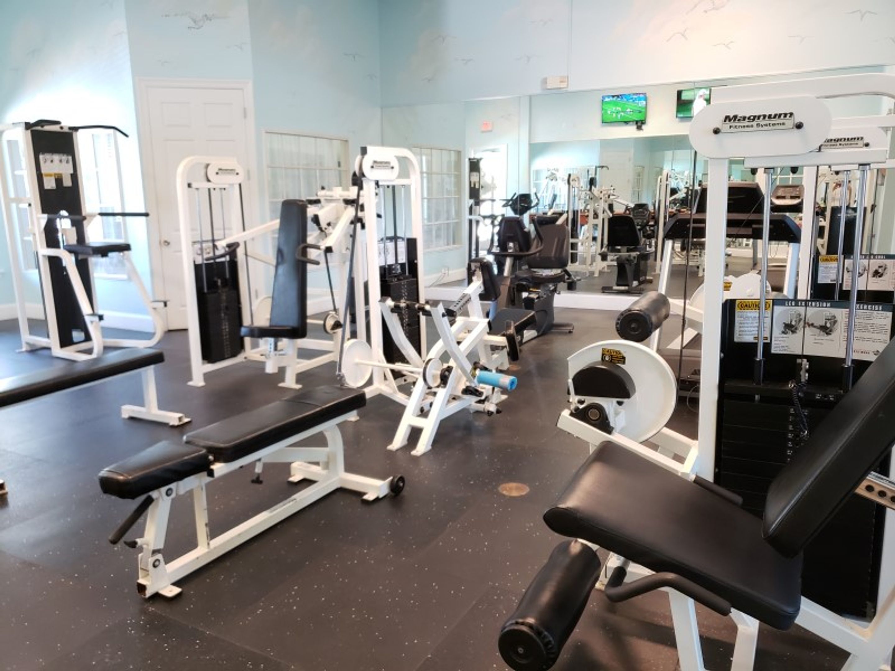 A gym with various exercise machines, benches, and a television screen on the wall.