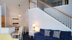 Un salon avec un canapé bleu, un fauteuil jaune, une table basse blanche, un coin repas avec une table et des chaises en bois, et un escalier avec des rampes blanches.