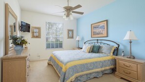 Ein Schlafzimmer mit Bett, Nachttischen, Kommode, Deckenventilator und Fenster mit Jalousien.