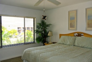 A bedroom with a bed, a ceiling fan, a window with blinds, a plant, and two framed pictures on the wall. - Fort Myers Beach Area 7327 - 302 (Fort Myers Beach)