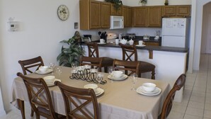 A dining area with a table set for six, surrounded by wooden chairs. A kitchen with wooden cabinets and a microwave is visible in the background. - High Gate Park 938 (Davenport)