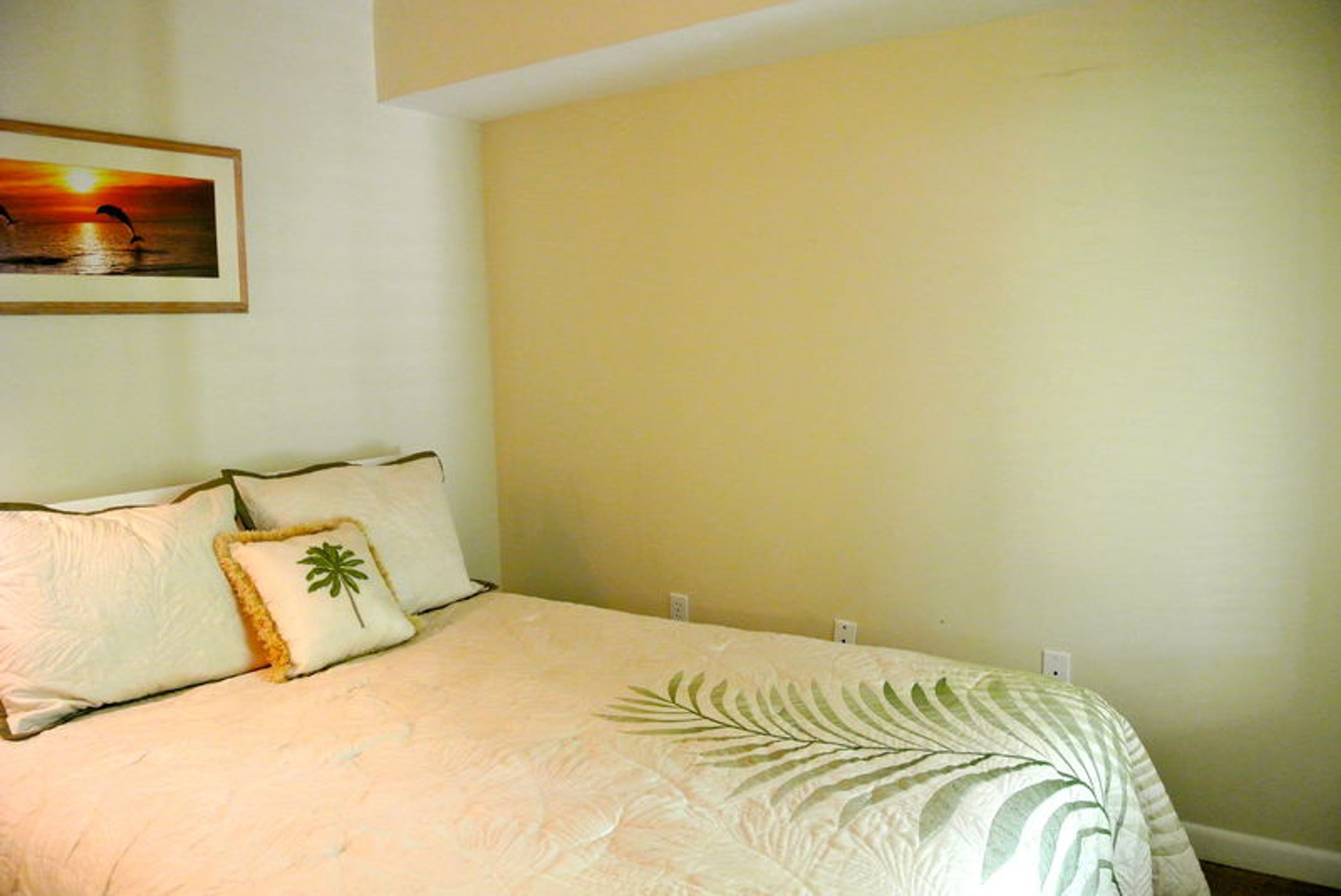 A bedroom with a bed, pillows, a framed picture on the wall, and a plain wall.