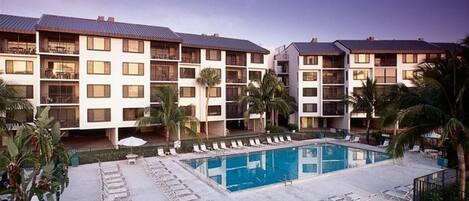 A hotel with a central swimming pool surrounded by lounge chairs and palm trees.