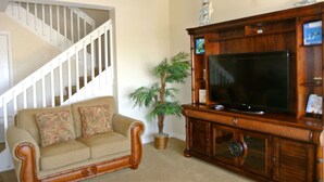 A living room with a beige sofa, a television on a wooden stand, a staircase, and a potted plant.