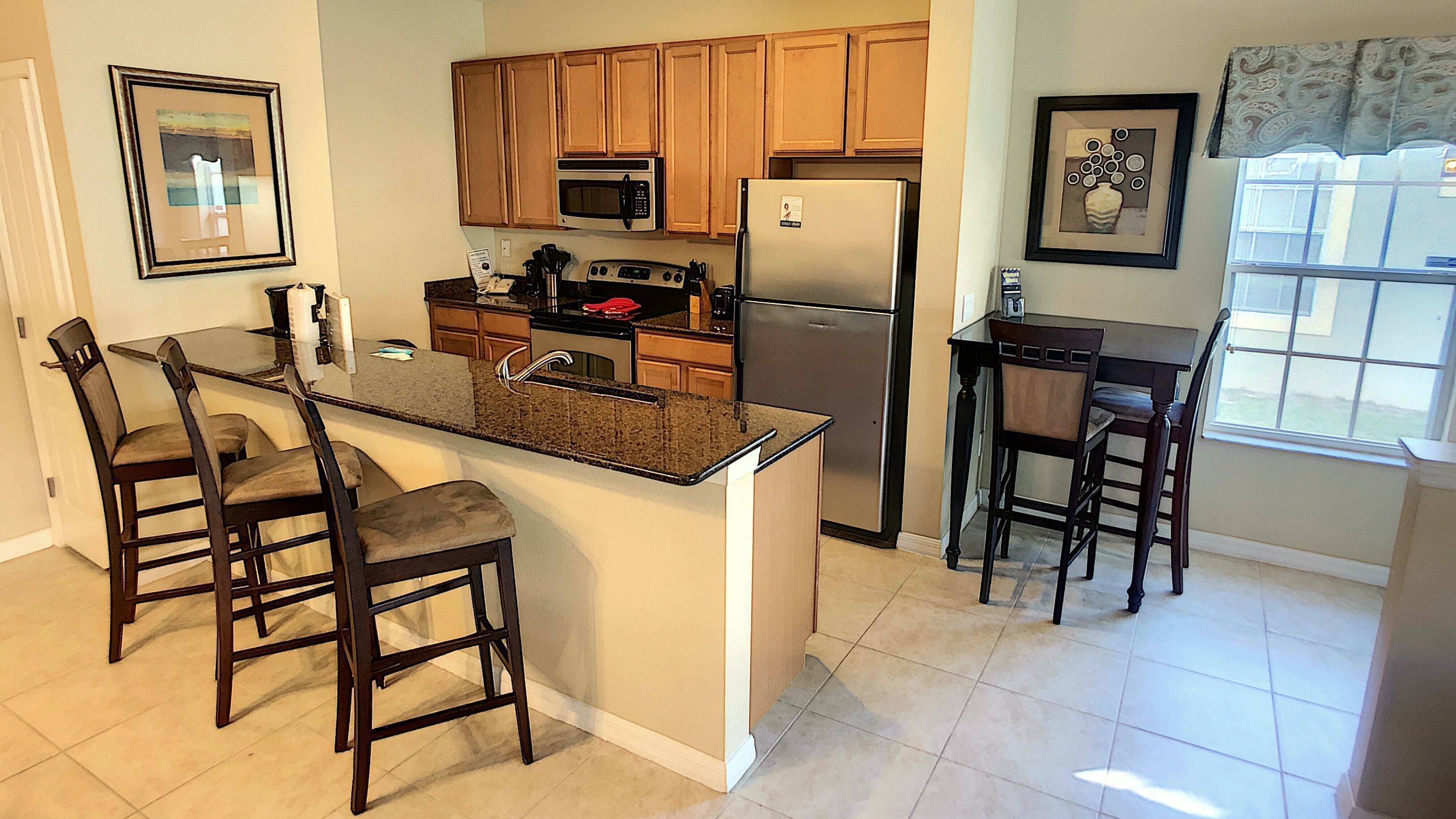 A kitchen with wooden cabinets, a stainless steel refrigerator, a microwave, and a granite countertop island with bar stools.