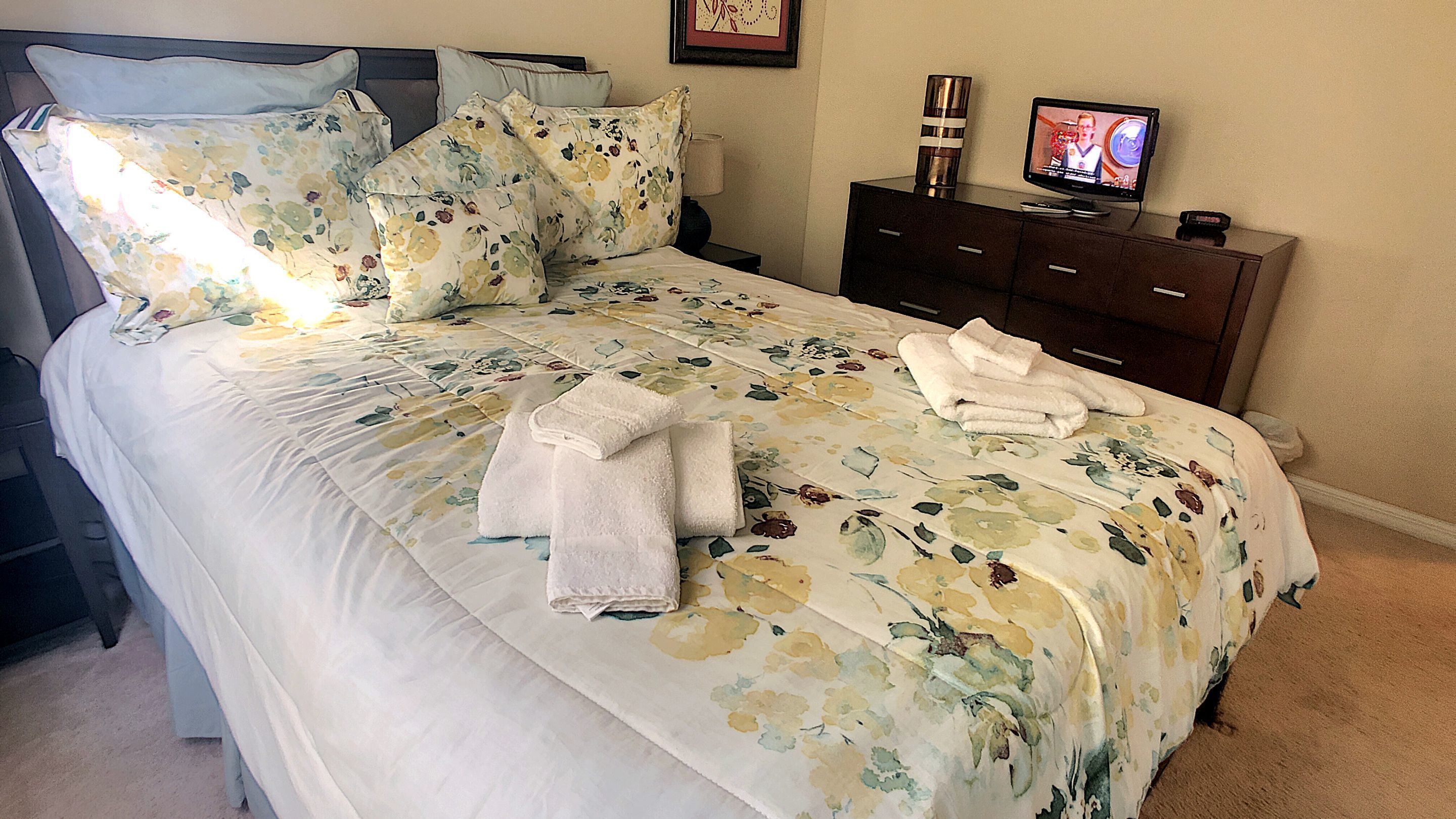 Una cama bien hecha con ropa de cama floral, un juego de toallas y un televisor sobre un aparador.