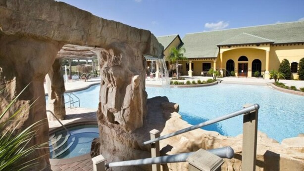 A pool area with a waterfall feature, a stone arch, and a building with a yellow facade and arched windows. - Paradise Palms 8965 Candy (Kissimmee)