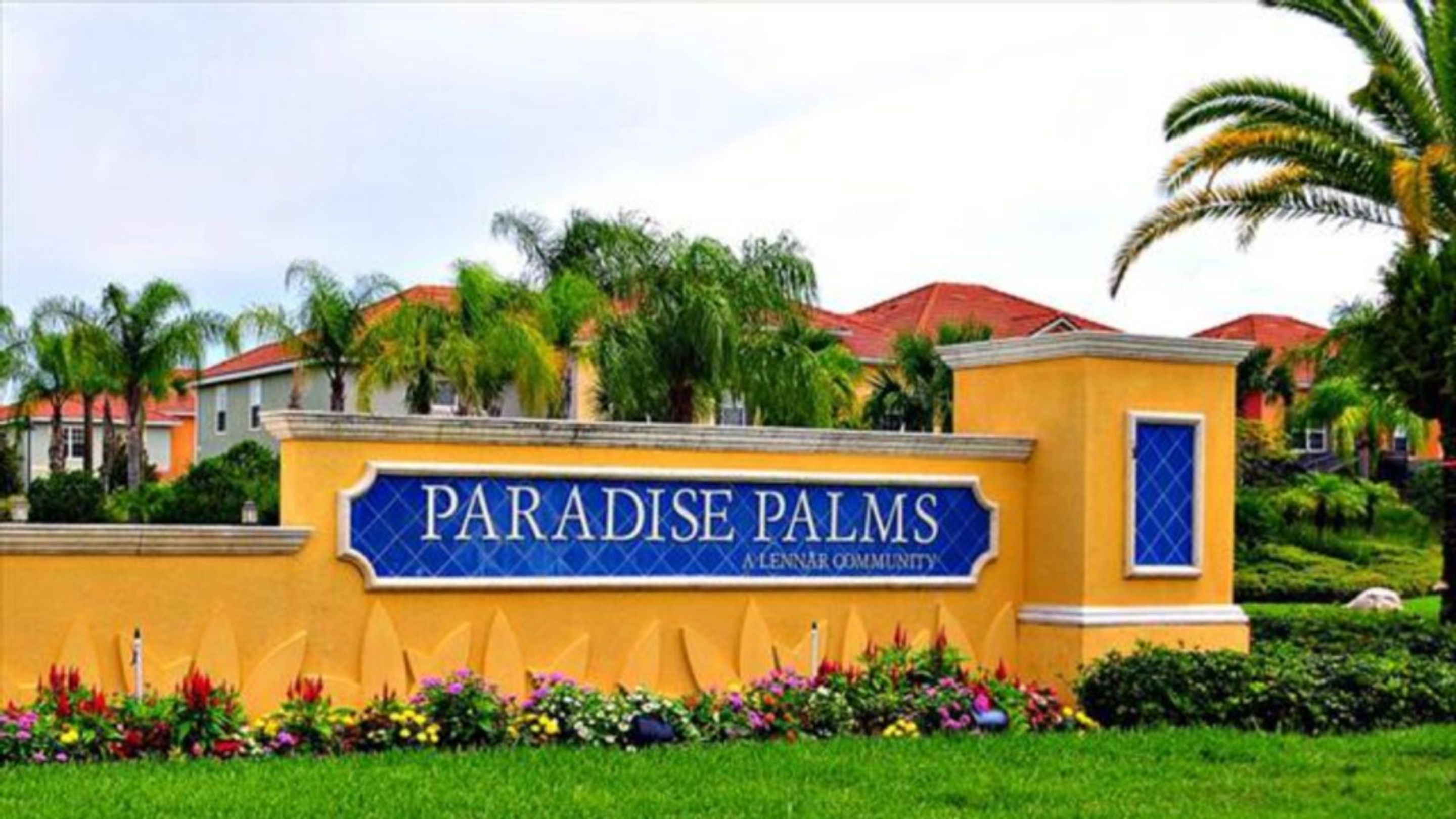 A sign for Paradise Palms, a Lennar community, with a yellow wall and a blue signboard.